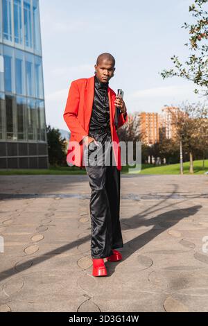 A young LGBTQ man confidently walks outdoors, sporting a stylish red jacket and holding a phone. The urban background adds to the vibrant and modern a Stock Photo