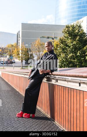 Young LGBTQ man in stylish black leather jacket and red boots leaning confidently against a bridge railing in an urban setting. Background of modern b Stock Photo