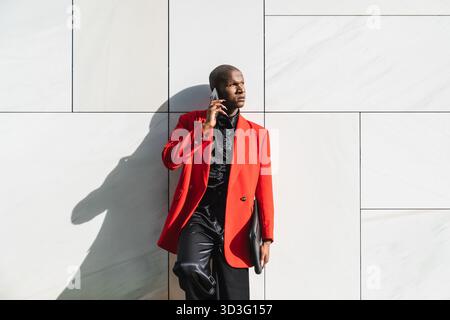 A confident young LGBTQ man stands against a modern tiled wall, wearing a striking red jacket. He is engaged in a phone conversation, exuding style an Stock Photo