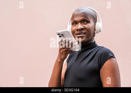 A young LGBTQ man stands confidently, smiling and interacting with his smartphone while wearing headphones against a neutral background, embracing con Stock Photo