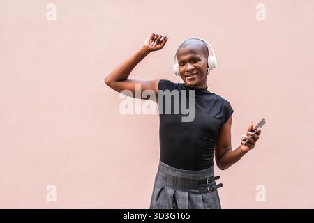 A young LGBTQ man joyfully listens to music on headphones, eyes closed in delight. Holding a smartphone, he dances in a relaxed manner against a neutr Stock Photo