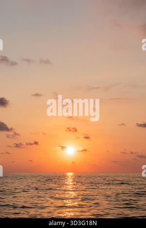 A breathtaking sunset over a tranquil sea, with the glowing sun reflecting on the water's surface. Soft clouds scatter across the sky, creating a sere Stock Photo