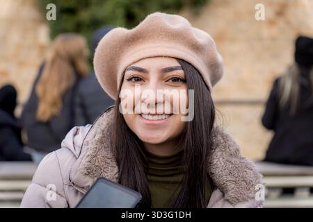 A young woman in Paris is smiling warmly, wearing a stylish beige beret and cozy jacket. The background features a charming cityscape that adds Parisi Stock Photo