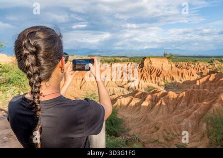 Back view of unrecognizable woman using a smartphone to capture the unique landscape of the Tatacoa Red Desert in Colombia, under a vast blue sky. Stock Photo