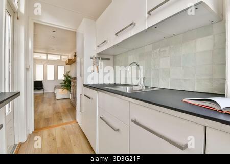 A sleek, modern kitchen featuring white cabinets, a black countertop, and a sink. The bright room opens into a cozy living space with wooden flooring Stock Photo
