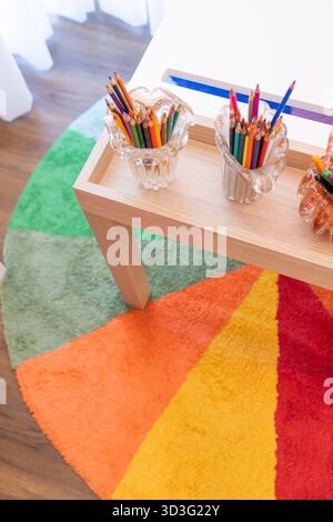 A vibrant study area for children featuring a rainbow-colored circular rug, colorful pencils in glass holders, and a wooden table, creating a playful Stock Photo