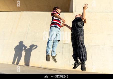 A joyful couple jumps in unison against a concrete backdrop, casting playful shadows in the bright sunlight. This image captures an expression of care Stock Photo