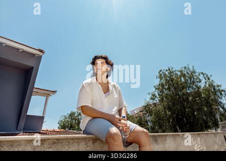 A young man sits outdoors holding a water bottle, enjoying a sunny day. Captured from below, the image emphasizes the bright blue sky and summer warmt Stock Photo