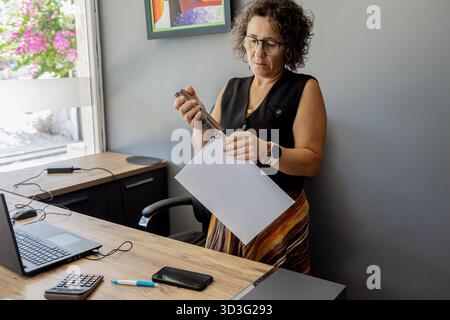 Woman standing at a wooden desk in a modern office, using a stapler to attach a stack of white papers. A laptop, smartphone, calculator, and pen are s Stock Photo