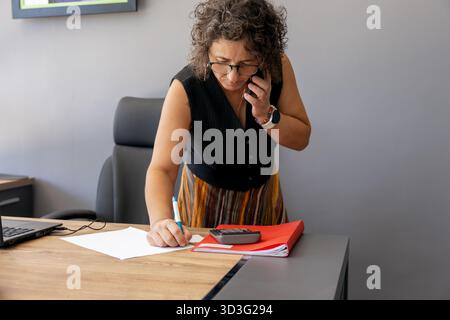 A professional woman in an office multitasks effectively, speaking on a cellphone while taking notes. Her desk is organized with a laptop and a calcul Stock Photo