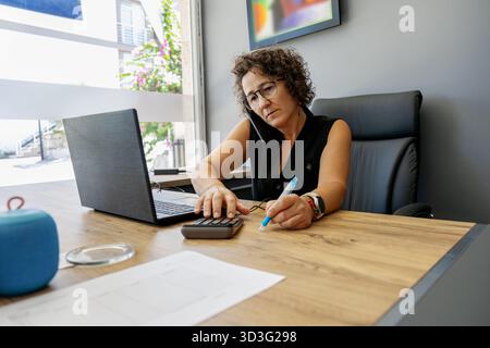 Businesswoman multitasking at a modern office desk. She is using a laptop, calculator, and phone while taking notes. A bright, productive work environ Stock Photo