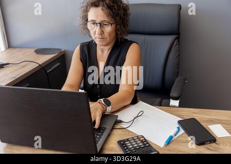 A woman focuses intently on her laptop in a modern office setting. She's surrounded by work tools, including a calculator, smartphone, and pen, portra Stock Photo