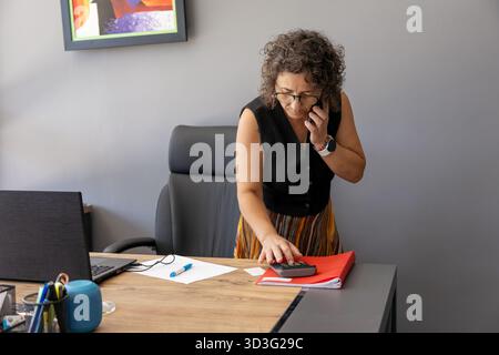 A businesswoman in a home office setting, multitasking by talking on the phone while using a calculator. She is surrounded by office supplies, a lapto Stock Photo