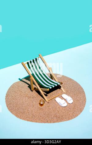 A green and white striped miniature deck chair and a small glass sit on a circle of sand, while white slippers rest beside it, set against a bright bl Stock Photo