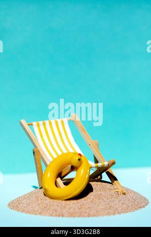 A wooden deck chair with yellow and white striped fabric sits on sandy beach. Beside it, a bright yellow inflatable ring is placed, against a clear bl Stock Photo