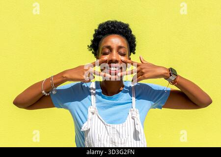 A cheerful black woman playfully poses with her fingers framing her face, standing in front of a vibrant yellow background. She wears bracelets and a Stock Photo