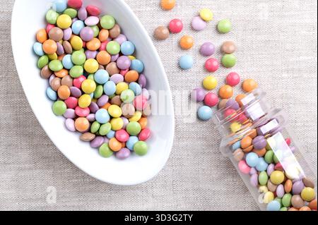 Candy coated mini chocolate lentils, in a white bowl, on linen. Small, sugar-coated chocolate dragees, with multi colored candy shell. Stock Photo
