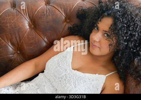 A woman with curly hair relaxes on a leather sofa, wearing a delicate white lace dress. Her serene expression embodies a moment of peaceful introspect Stock Photo