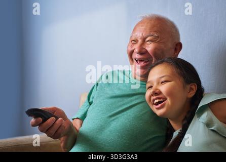 A joyful senior man shares a heartwarming moment with his granddaughter as they sit on the sofa and watch TV. Both are smiling, enjoying their time to Stock Photo