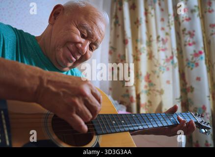 An elderly man plays an acoustic guitar at home, joyfully strumming the strings. He is seated comfortably indoors, showcasing a relaxed and content ex Stock Photo