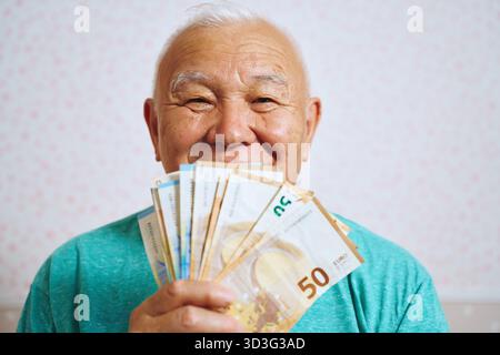 A cheerful elderly man at home, smiling as he holds several euro bills in front of him. The image captures a joyful moment of financial success and co Stock Photo