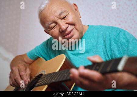 An elderly man is comfortably at home playing an acoustic guitar, smiling as he embraces the music. His joyful expression reflects a deep love for mus Stock Photo