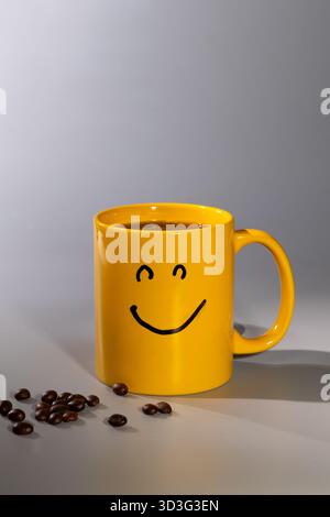A cheerful yellow mug featuring a smiley face sits on a clean surface with coffee inside. Coffee beans are scattered nearby, enhancing the positive morning vibe. Stock Photo