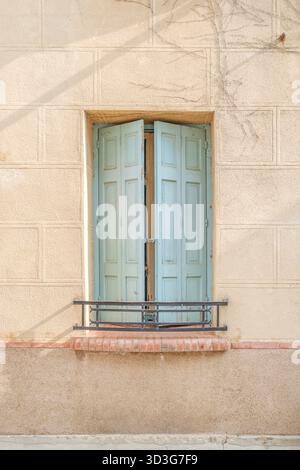 Single window with light blue wooden shutters slightly ajar, set in a neutral stucco wall with a red brick ledge and wrought iron railing Stock Photo