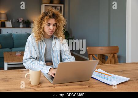 Young woman working from home on laptop, casual striped shirt and coffee nearby, reviewing charts and typing with calm focus remote work productivity Stock Photo
