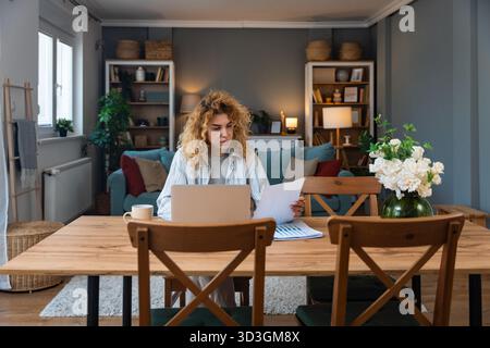 Young woman working from home on laptop, casual striped shirt and coffee nearby, reviewing charts and typing with calm focus remote work productivity Stock Photo