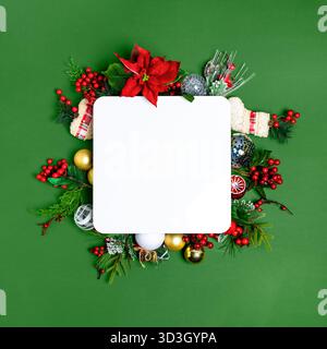 A festive Christmas flat lay with a blank white mockup card, surrounded by poinsettia, berries, and baubles on a green background Stock Photo