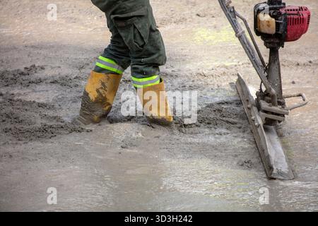 Worker in yellow boots leveling wet concrete on construction site outdoors, using vibrating screed under daylight. Stock Photo