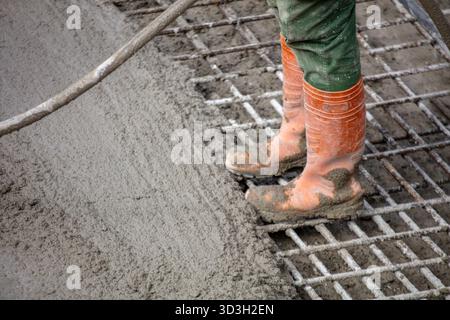Construction worker pouring wet concrete over steel bars, wearing orange boots and green pants, seen from above on a cloudy day. Stock Photo