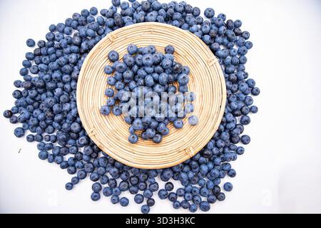 Fresh blueberries on round wicker plate surrounded by scattered berries on white background Stock Photo