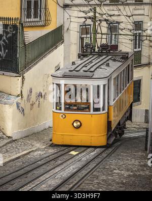 View of a vibrant yellow funicular ascends the steep, cobbled street, framed by weathered buildings with intricate wrought-iron balconies, Lisbon, Lis Stock Photo