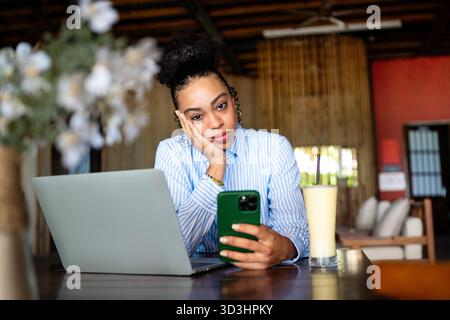 African american woman with a phone and laptop in a cozy space feeling unengaged while waiting Stock Photo