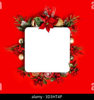 A festive Christmas flat lay with a blank white mockup card, surrounded by poinsettia, berries, and baubles on a red background. Stock Photo