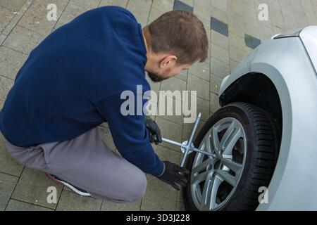 Man changing car tire using electric drill and jack on city sidewalk. DIY auto repair, maintenance, seasonal tire change, mechanical work, vehicle Stock Photo