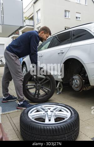 Man changing car tire using electric drill and jack on city sidewalk. DIY auto repair, maintenance, seasonal tire change, mechanical work, vehicle Stock Photo