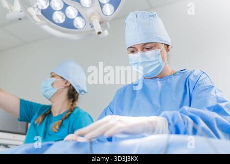 Two medical professionals in surgical attire focus on a procedure in a bright operating room, ensuring patient care. Stock Photo