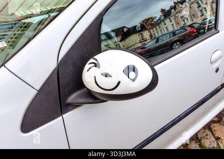 A funny winking face illustration hand-painted on the external mirror of a Citroen C1 car - Tours, Indre-et-Loire (37), France. Stock Photo