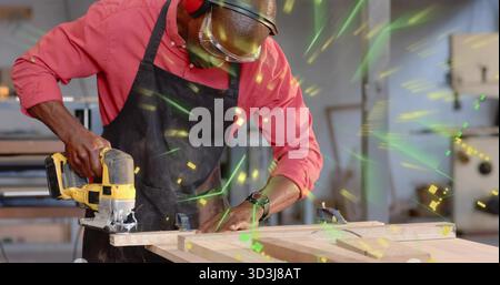 Cutting wooden plank with handheld jigsaw saw in woodworking shop, wearing apron and safety goggles Stock Photo
