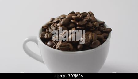 Displaying white ceramic coffee mug holding dark roasted coffee beans on white surface Stock Photo