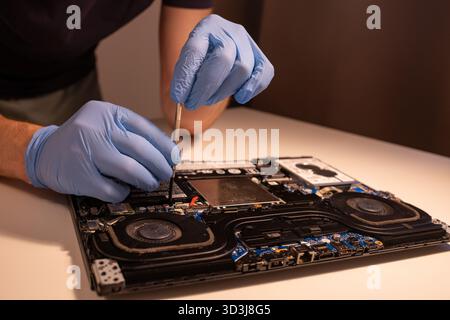 Technician wearing blue gloves using a screwdriver to repair or assemble a laptop’s internal components. Close-up of computer hardware maintenance Stock Photo