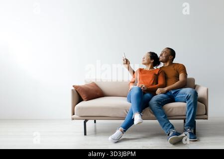 Split system. Cheerful young black couple opening air conditioner with remote Stock Photo