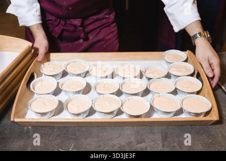 Food, A chef in a burgundy apron holding a tray filled with foil cups of light brown cupcake batter, ready for baking in the oven Stock Photo