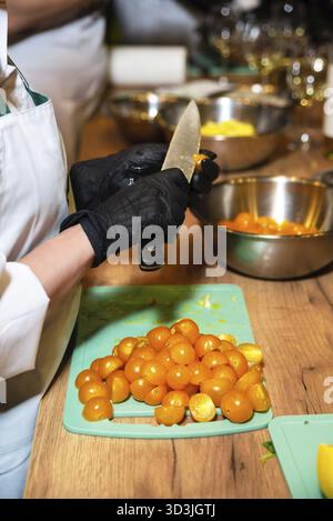 Food, Close-up of a person wearing black gloves cutting fresh yellow cherry tomatoes on a chopping board during food prep Stock Photo