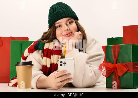 A young woman smiles while holding her phone amidst colorful Christmas presents and warm drinks. Stock Photo