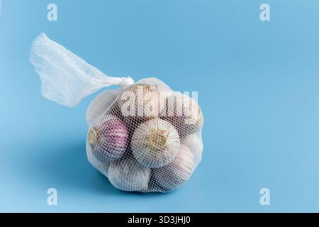 Small garlic bulbs in a mesh bag on a solid blue background. Stock Photo