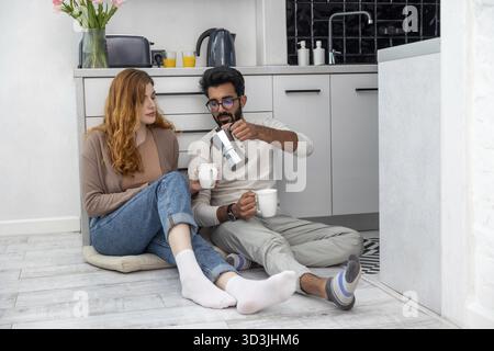 Wife and husband sitting on floor in kitchen and talking, drinking morning coffeee at home Stock Photo
